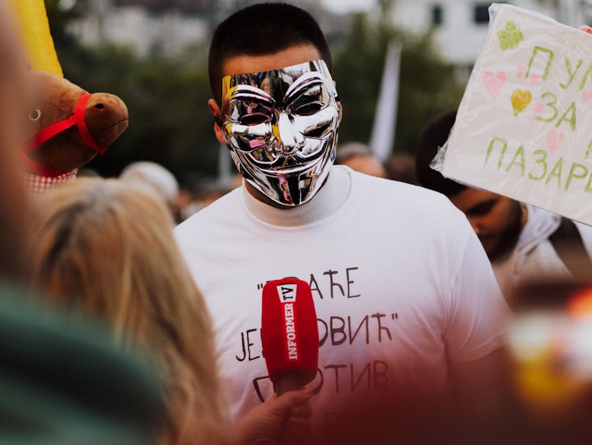 A person in an Anonymous mask at a protest in Belgrade, Serbia, holding a sign and microphone.