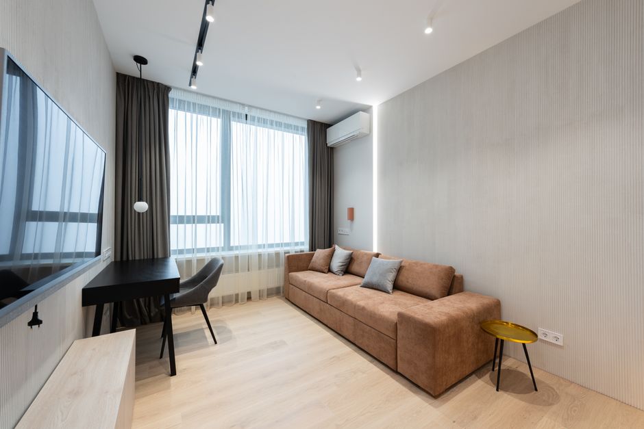 Interior of modern living room with sofa with pillows and TV on wall next to table with chair and curtains on window