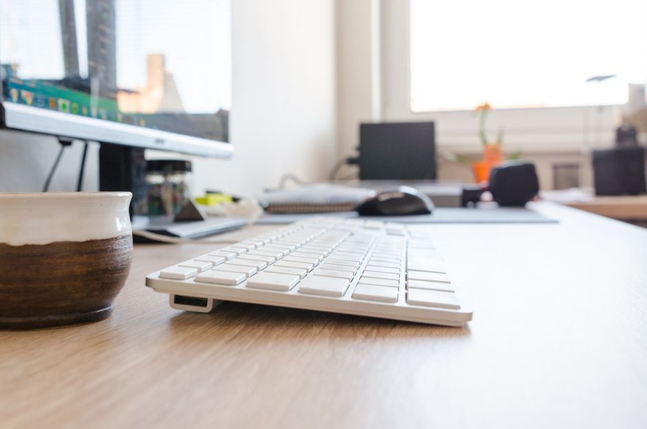 Minimalist office desk setup featuring keyboard, monitor, and coffee cup with natural lighting.