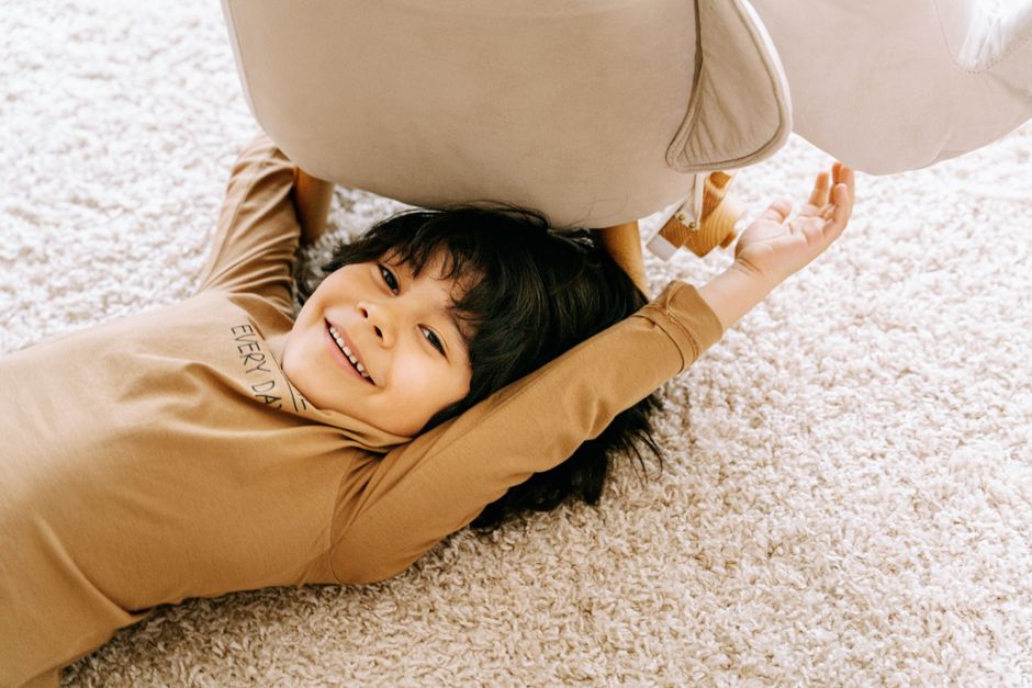 Smiling child lying under a chair on a plush carpet, enjoying a moment of playful relaxation.