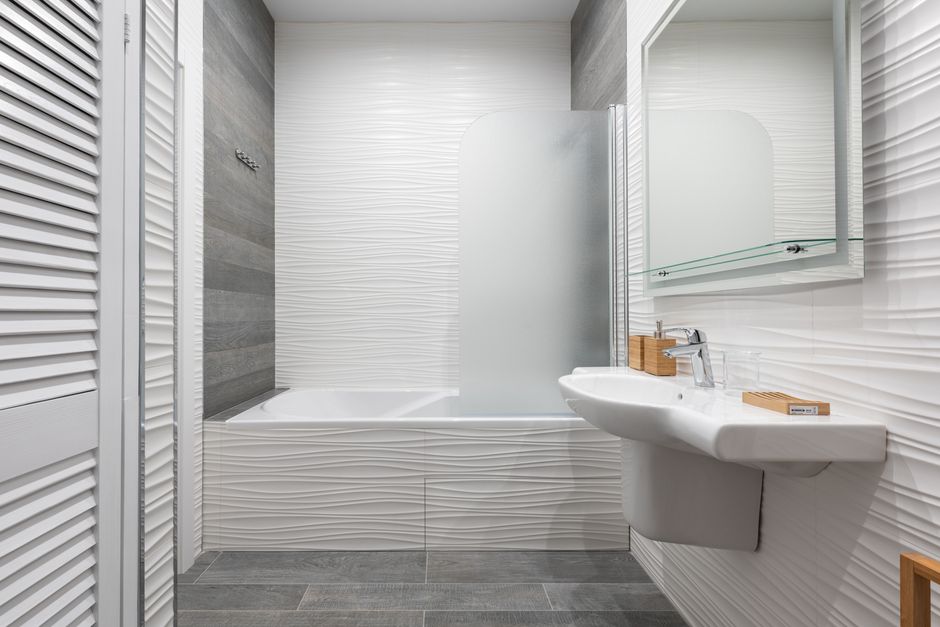 Washbasin with toiletry products and soap dish under mirror against bath and ribbed wall in light bathroom