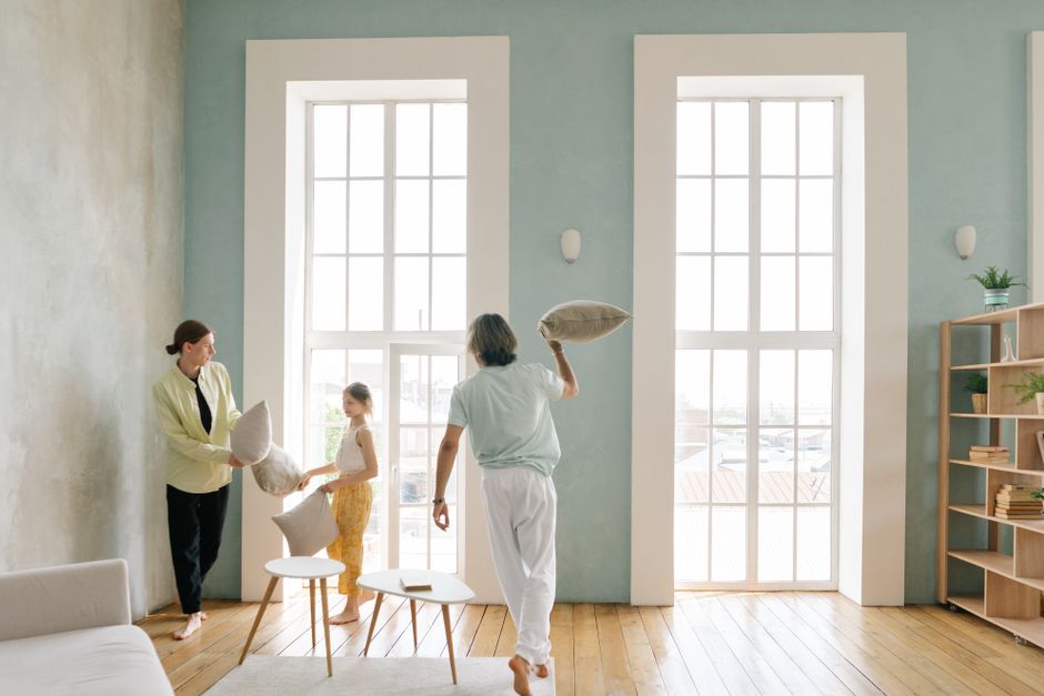 Playful family pillow fight in a bright modern living room with large windows.