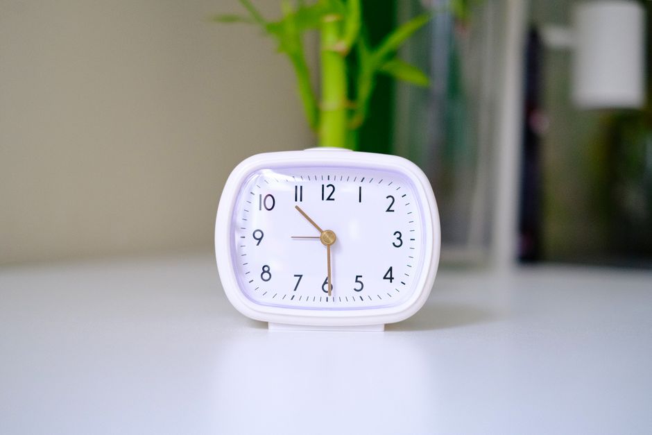 A minimalist white analogue clock placed on a clean desk with a blurred plant in the background.