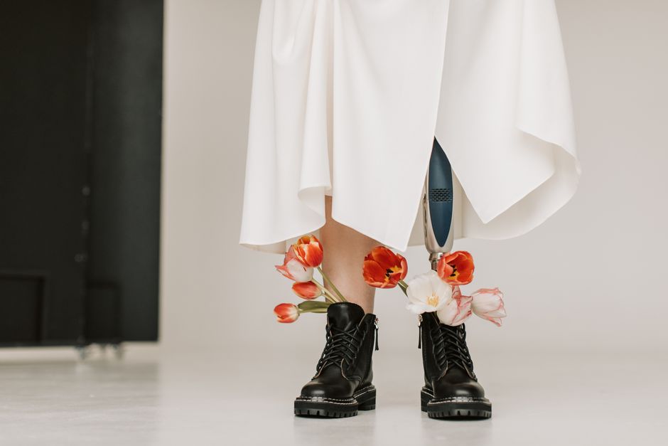 Close-up of a person wearing a prosthetic leg, black boots, and a white dress adorned with tulips.