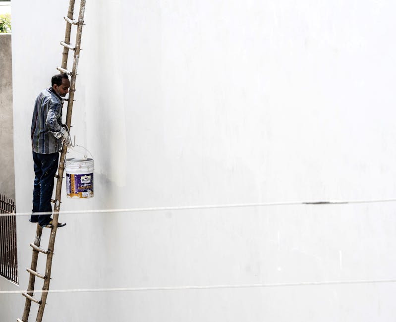 A worker on a ladder paints a large white wall outdoors, showcasing construction work.