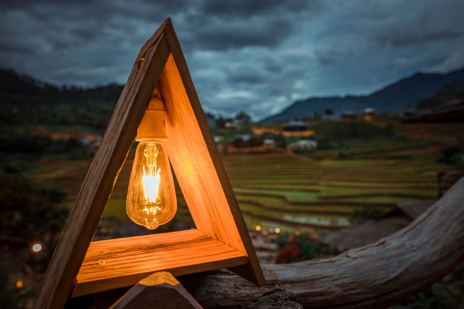 A glowing light bulb in a triangular wooden fixture with scenic rice terraces in the background under cloudy skies.
