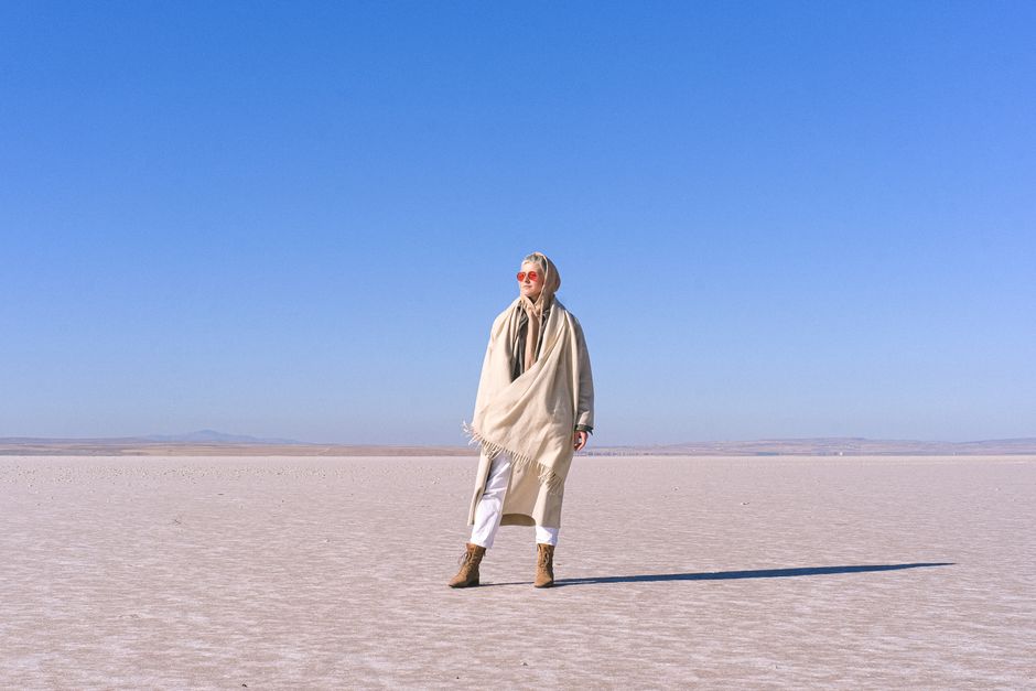 A stylish woman stands in a vast desert landscape, wrapped in a scarf and wearing sunglasses.
