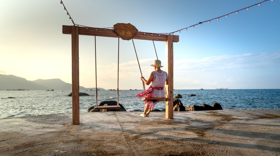 A woman sits on a swing overlooking a calm ocean, under a clear blue sky, conveying peace and relaxation.