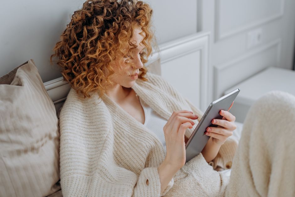 A woman relaxing on a bed, using a digital tablet while wearing a cozy knitted sweater.