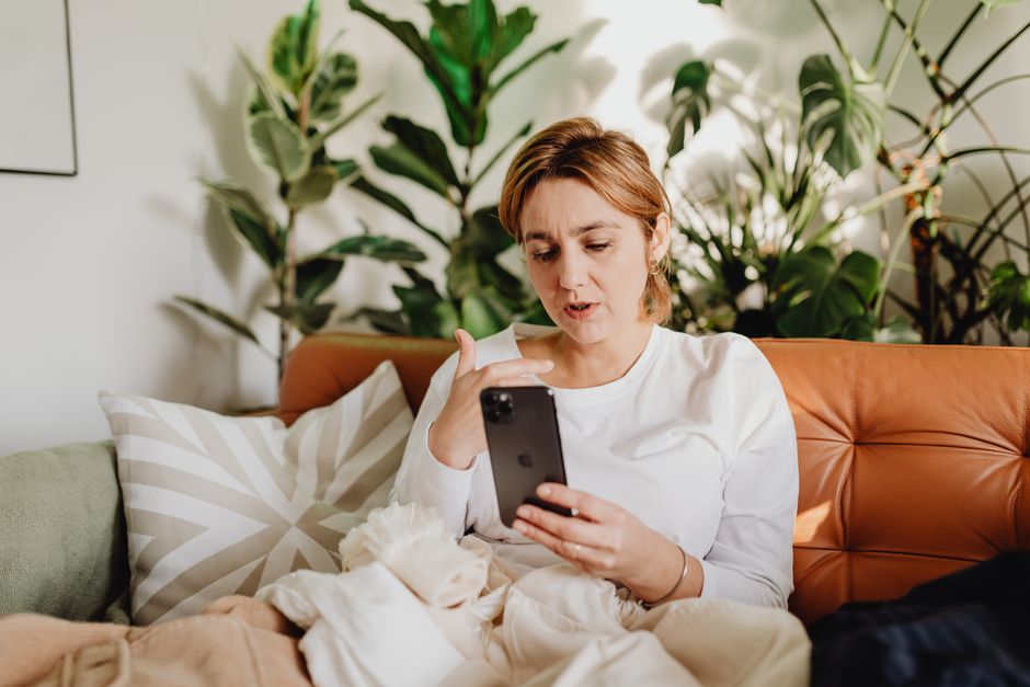 Woman in white shirt using smartphone on sofa surrounded by plants.