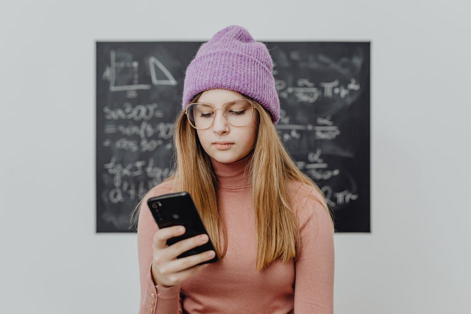 Teenager wearing a beanie and eyeglasses using a smartphone against a blackboard.
