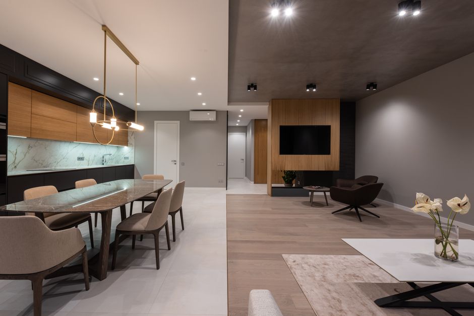 Modern apartment interior with dining table and chairs under pendant lamp next to open kitchen and armchairs near TV on wall