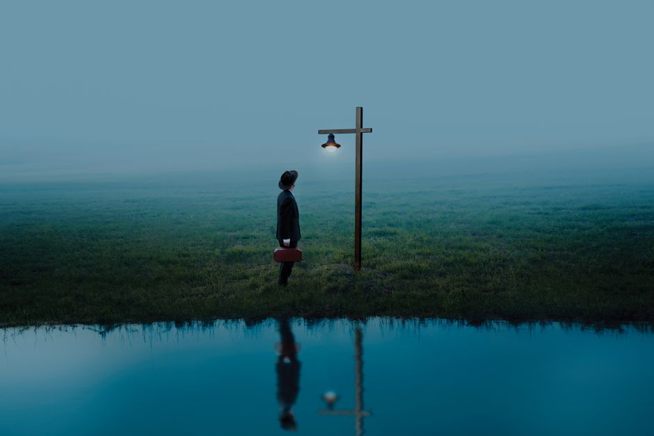A solitary figure stands under a light post by water, reflecting solitude and hope.